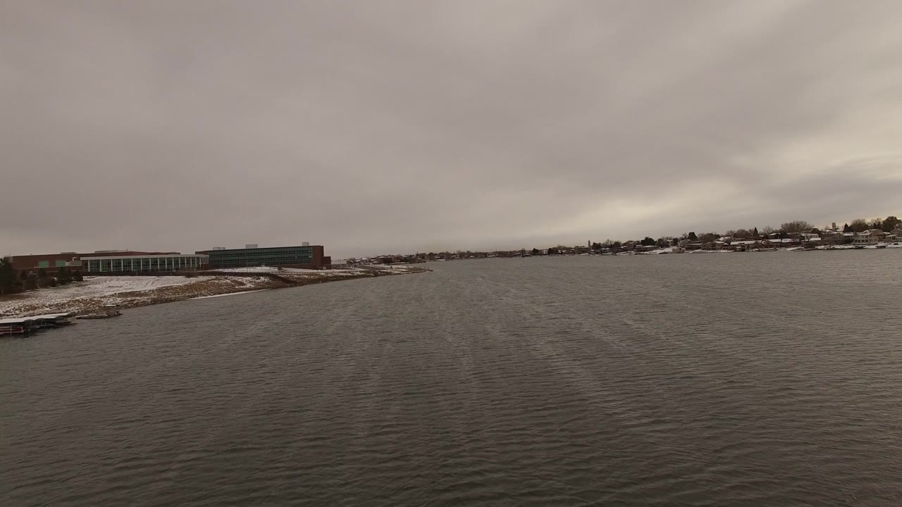 At 0:00, a wide elevated view in Jefferson, Colorado shows rippled water, a snow-dusted shoreline with low buildings on the left, distant houses across the bay, and no visible people.
