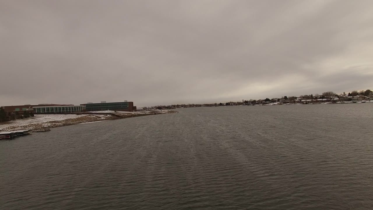 At 0:01, the Jefferson, Colorado bay remains centered, with dark water in the foreground, a small dock near the snowy left shoreline, low buildings, distant homes, and no visible people.
