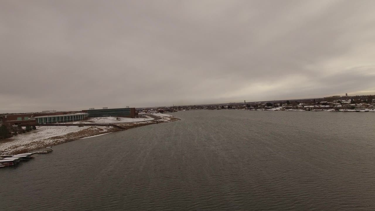 Aerial view over dark water beside snowy shoreline and buildings.