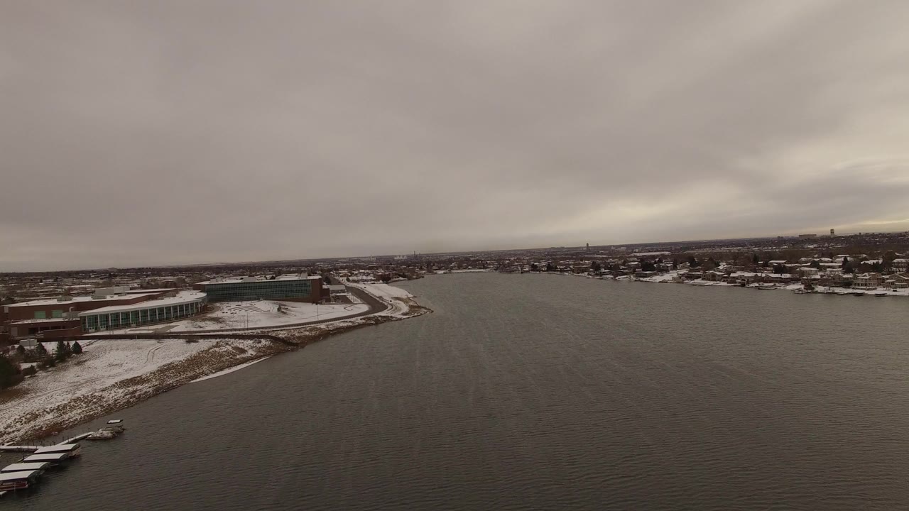 Aerial view over dark water toward a snowy shoreline and low buildings