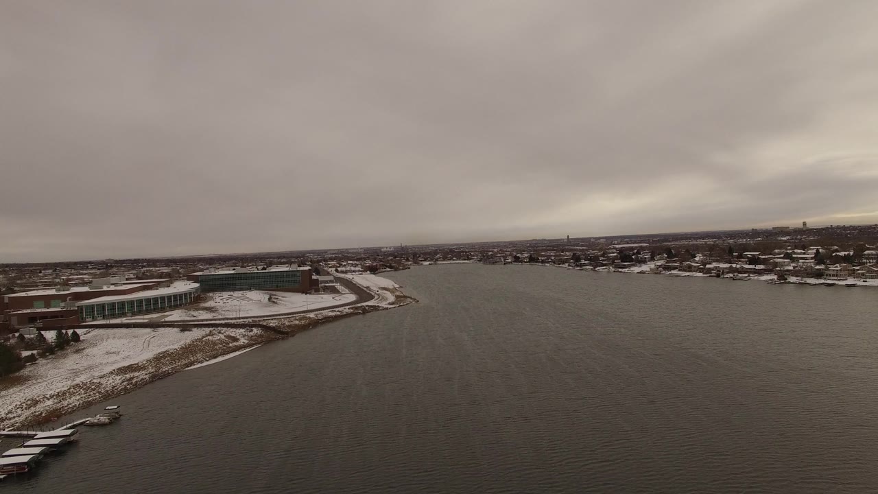 Aerial view in Jefferson, Colorado, looking across open water toward a curved snowy point, shoreline buildings, distant neighborhoods, low horizon, and no visible people.