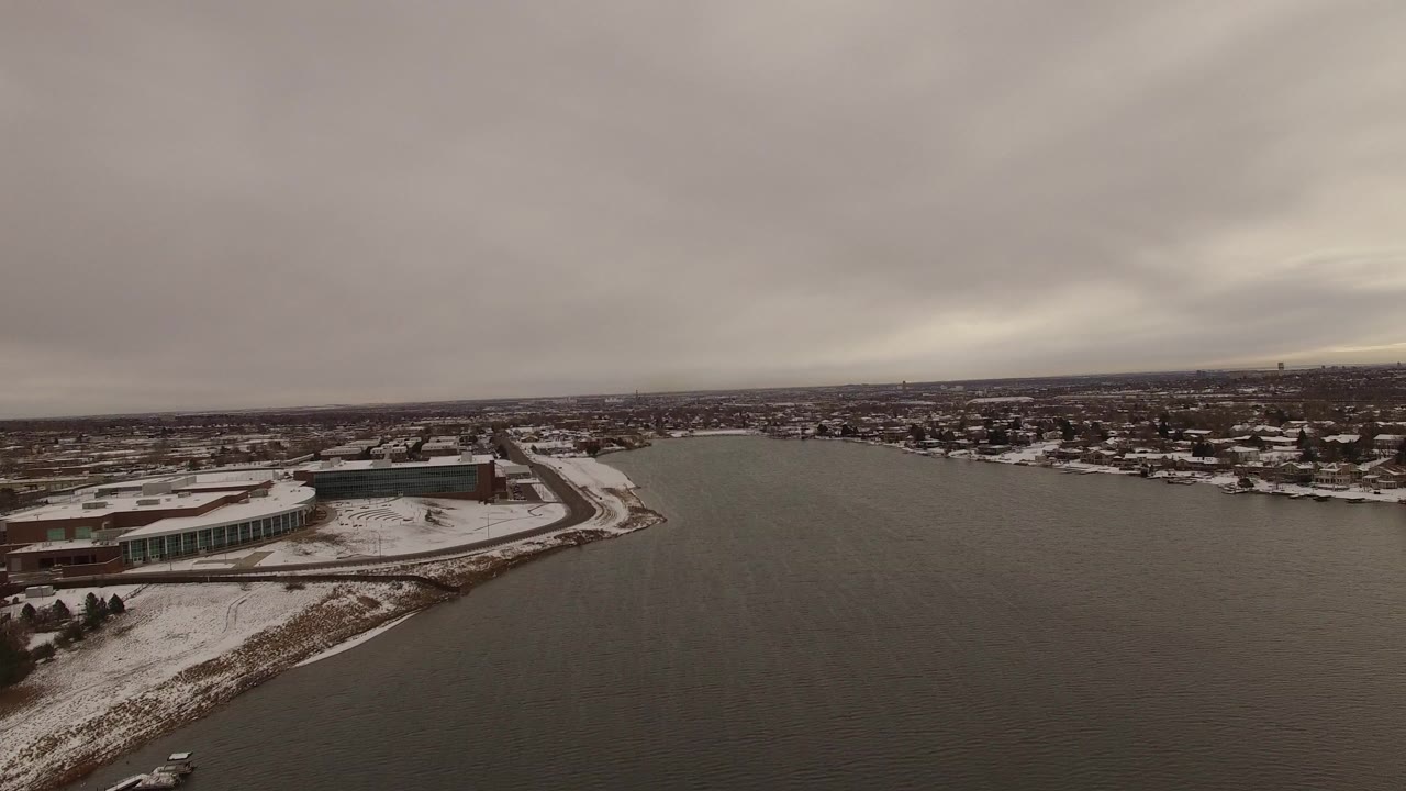 Aerial view of a snow-lined lake and shoreline buildings under gray clouds.