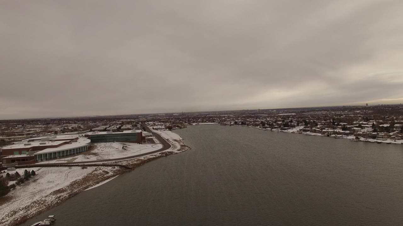 Aerial view in Jefferson, Colorado with the large shoreline building slightly more centered, dark water at right, snowy ground at left, town blocks beyond, and no visible people.