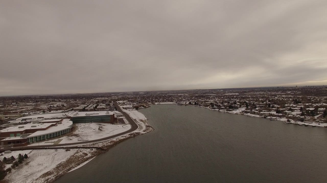 Snowy shoreline cityscape beside dark water under gray clouds.