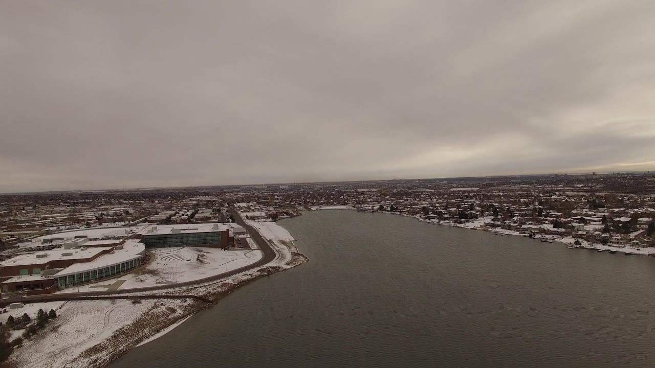 Aerial frame over Jefferson County, Colorado, with the water occupying the right foreground, snow-lined roads and buildings on the left, distant neighborhoods, and no visible people.
