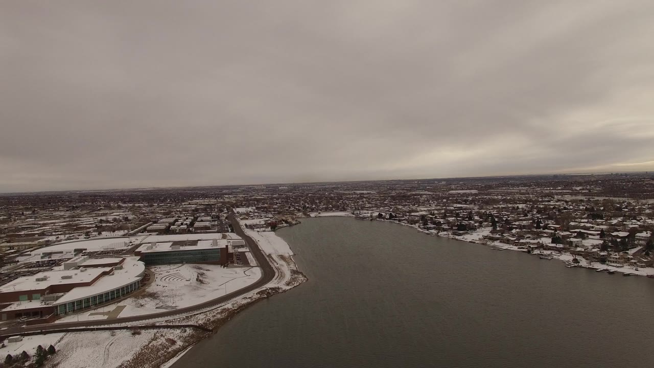 Aerial frame in Jefferson County, Colorado with the waterway centered, a snowy commercial-looking shoreline at left, residential blocks beyond, and no visible people.