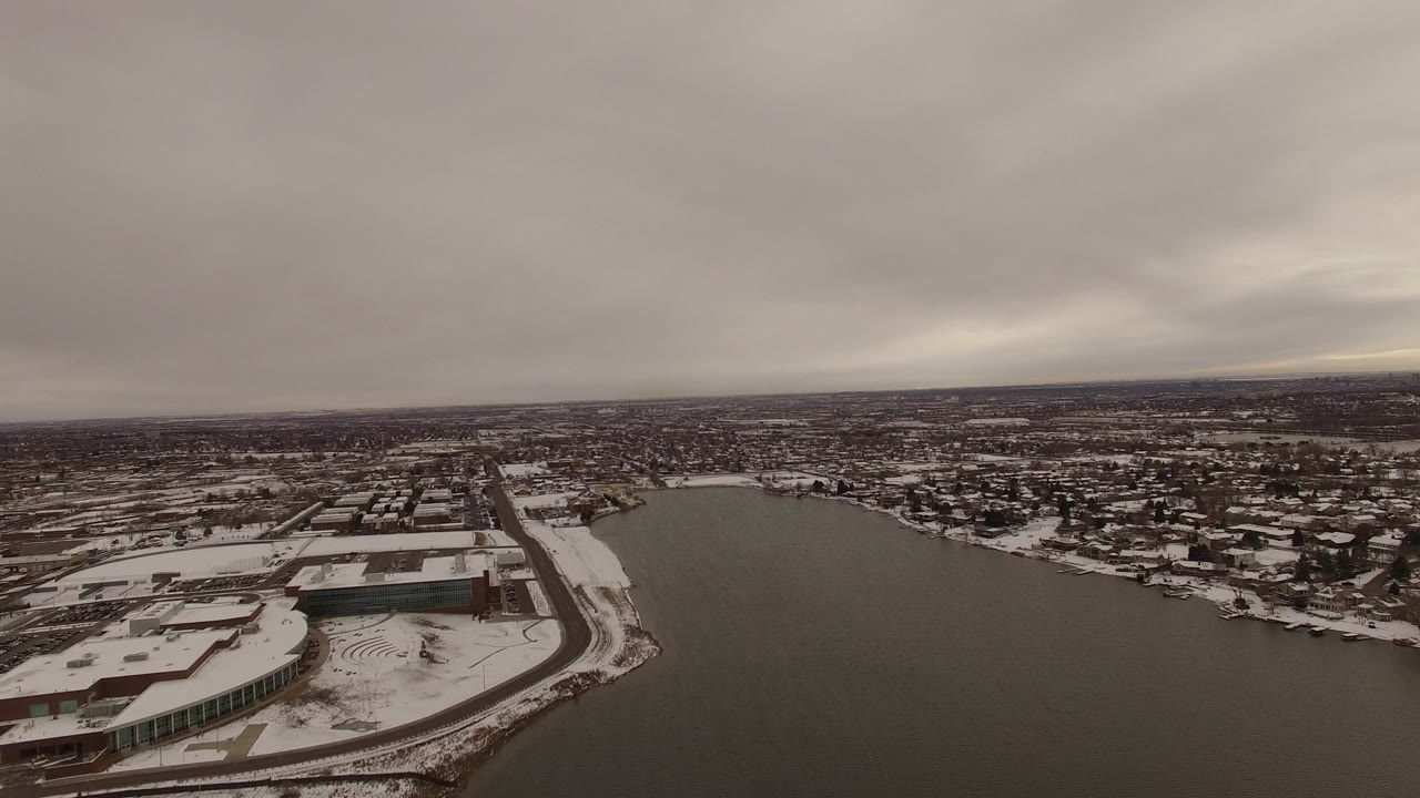 Opening aerial frame in Jefferson County, Colorado showing dark water beside a snow-covered shoreline, low buildings, distant neighborhoods, heavy cloud cover, and no visible people.