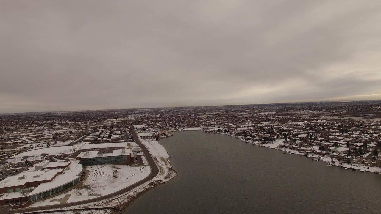 Aerial frame in Jefferson County, Colorado with the water shifted more centrally, snowy waterfront structures at lower left, dense residential blocks beyond, gray sky, and no visible people.