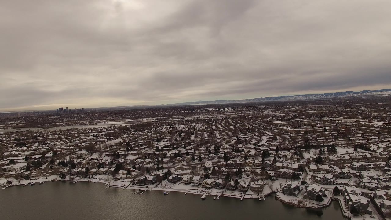 Snowy lakeside neighborhood under gray clouds with distant city skyline.