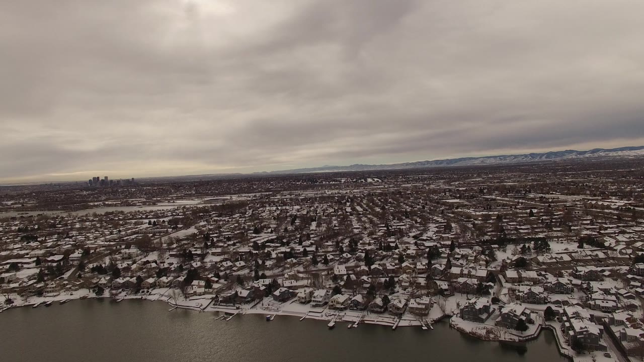The Jefferson County, Colorado aerial view centers more on dense snow-dusted homes, shoreline docks, dark water at the bottom edge, low gray clouds, distant mountains, and no discernible people.