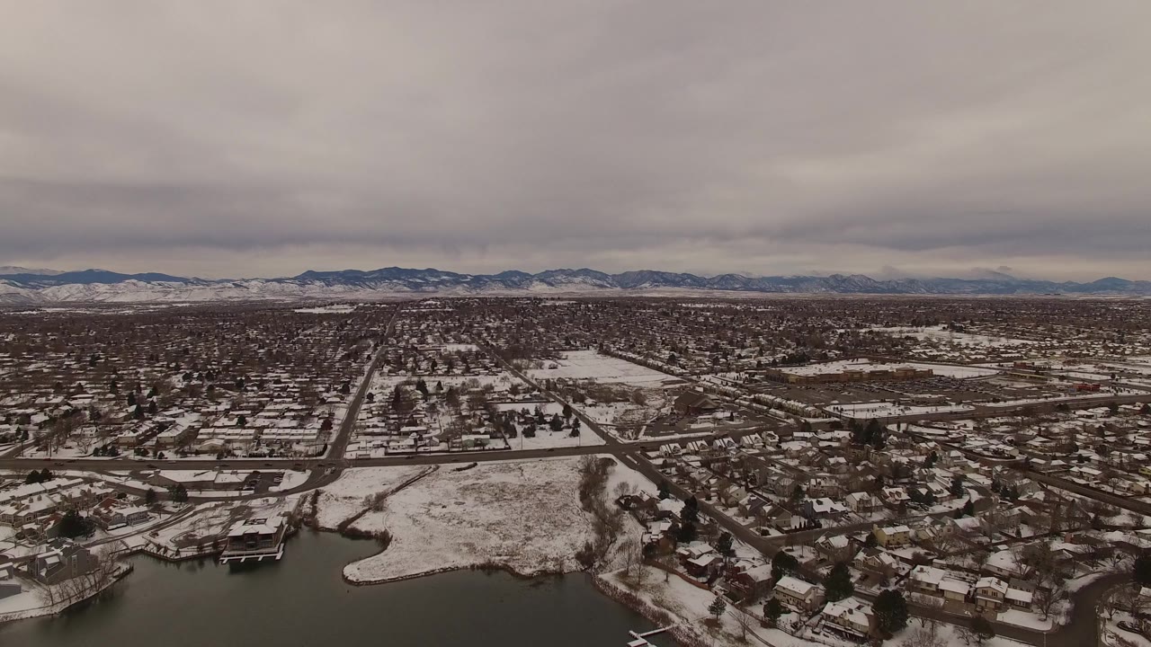 Jefferson County, Colorado aerial view with snowy homes and streets spreading across the frame, open white fields near the center, distant low mountains, heavy cloud cover, and no visible people.