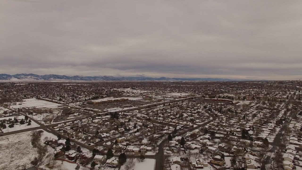 Second aerial frame in Jefferson County, Colorado looks across a snow-dusted suburban grid toward low mountains beneath heavy gray clouds, with no visible people.