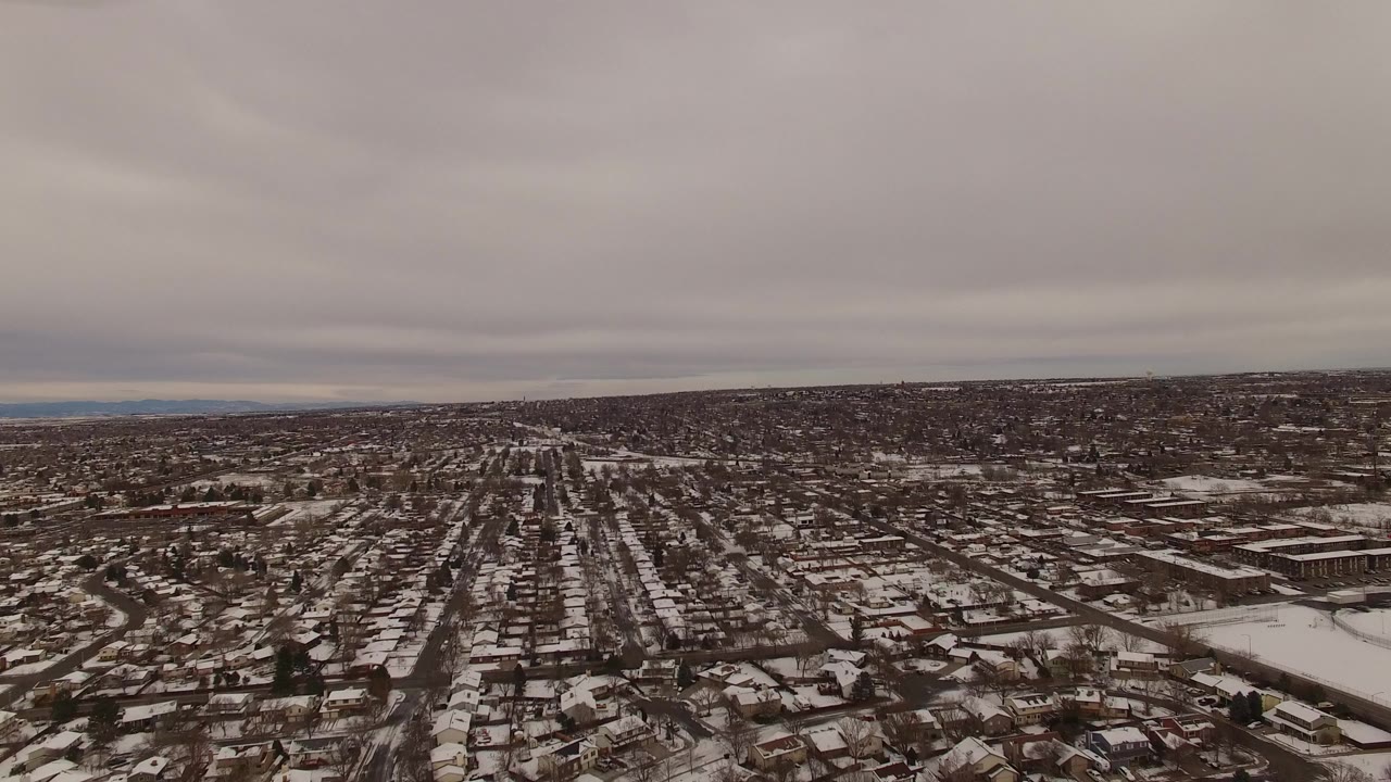 Wide aerial view over Jefferson County, Colorado residential blocks with snow on roofs and roads, distant hills on the horizon, and no visible people.