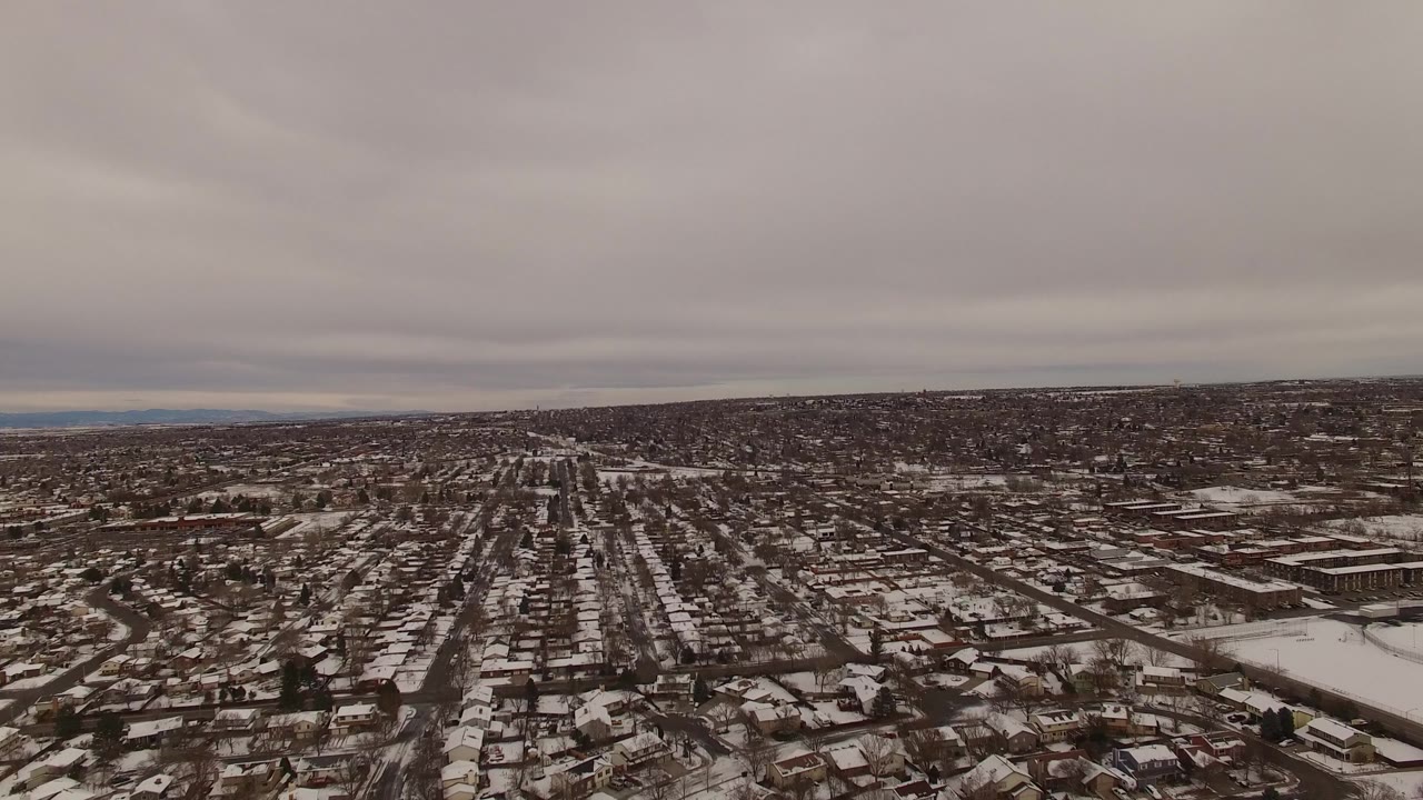 High aerial frame in Jefferson County, Colorado looking down a long road corridor through snow-dusted houses, with a broad gray cloud layer and no visible people.