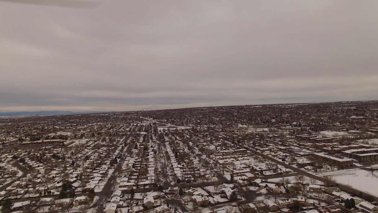 Aerial frame over Jefferson County, Colorado, with a straight street grid running toward the horizon, patchy snow on homes and yards, and no visible people.