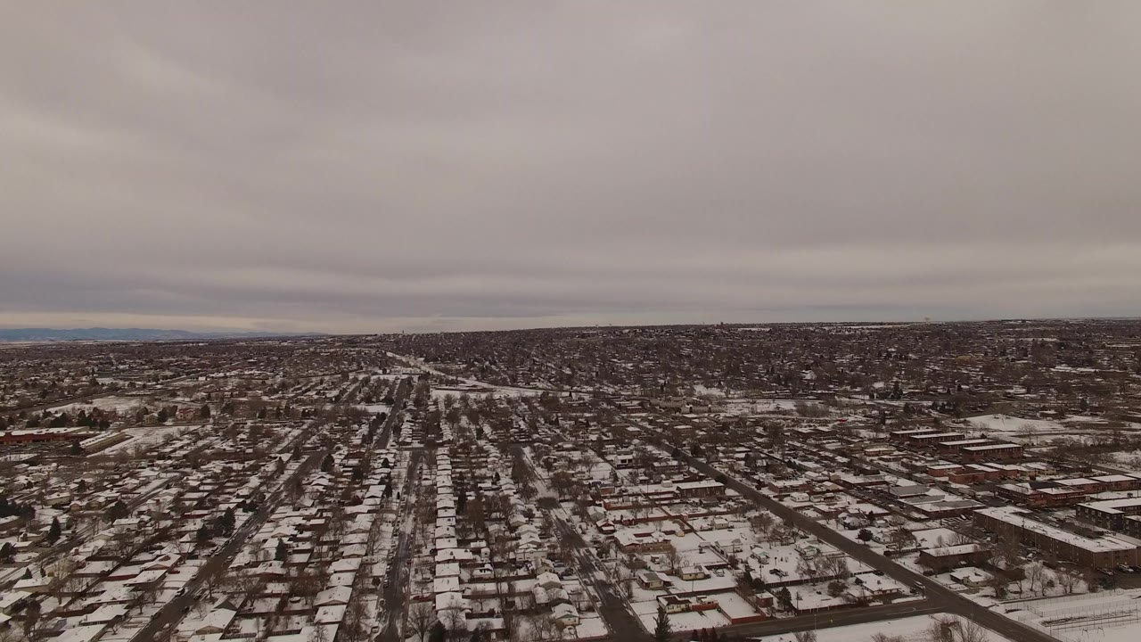 High aerial view in Jefferson County, Colorado with rows of snow-dusted homes, intersecting streets, scattered cars, and a low gray cloud ceiling with no visible people.