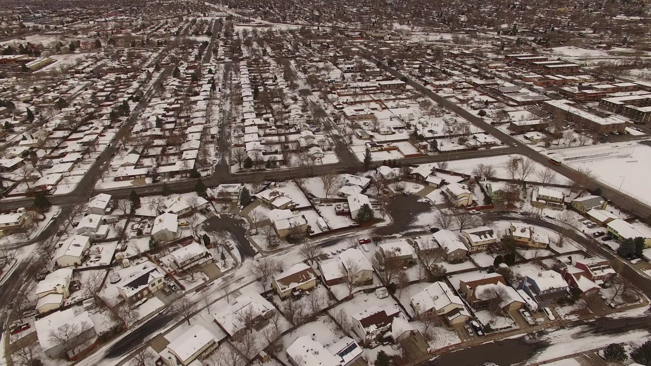 High aerial view over Jefferson County, Colorado homes with snow on rooftops and lawns, curving residential streets in front, long blocks in the distance, and no visible people.