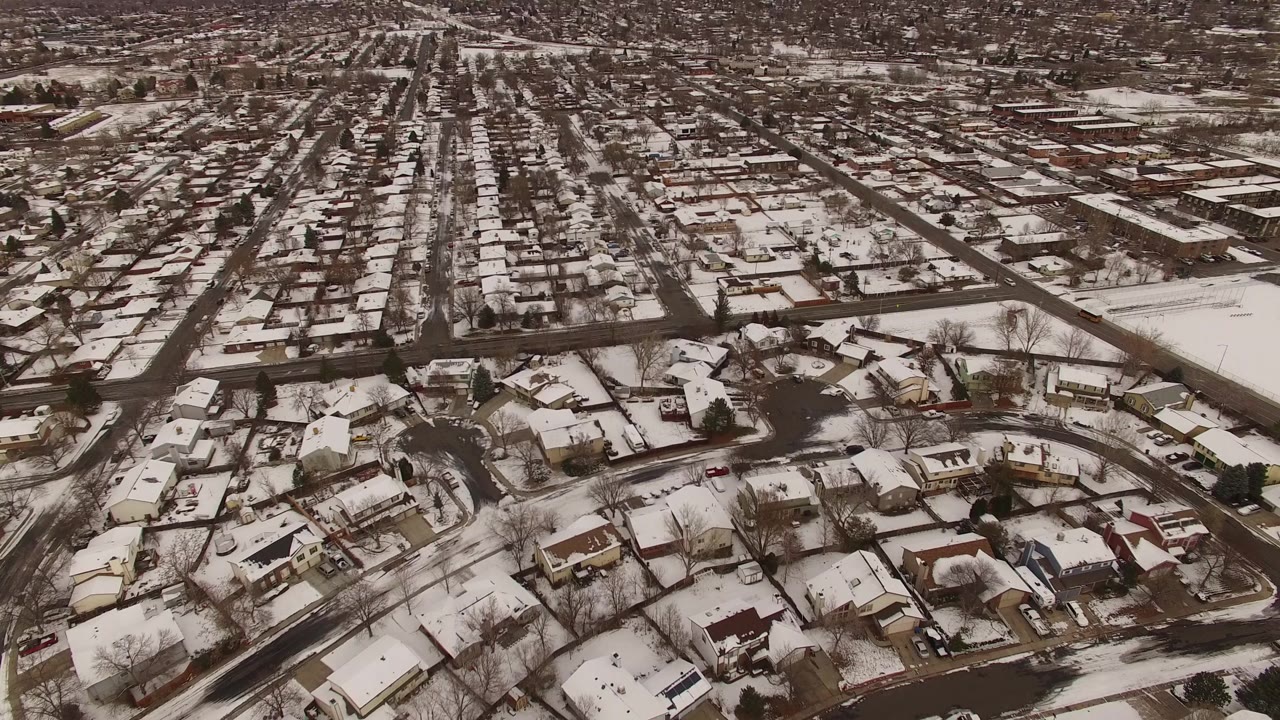 Aerial frame over Jefferson County, Colorado showing a snowy subdivision with dark roads, white roofs, bare trees, and a long straight street corridor extending into the background with no visible people.
