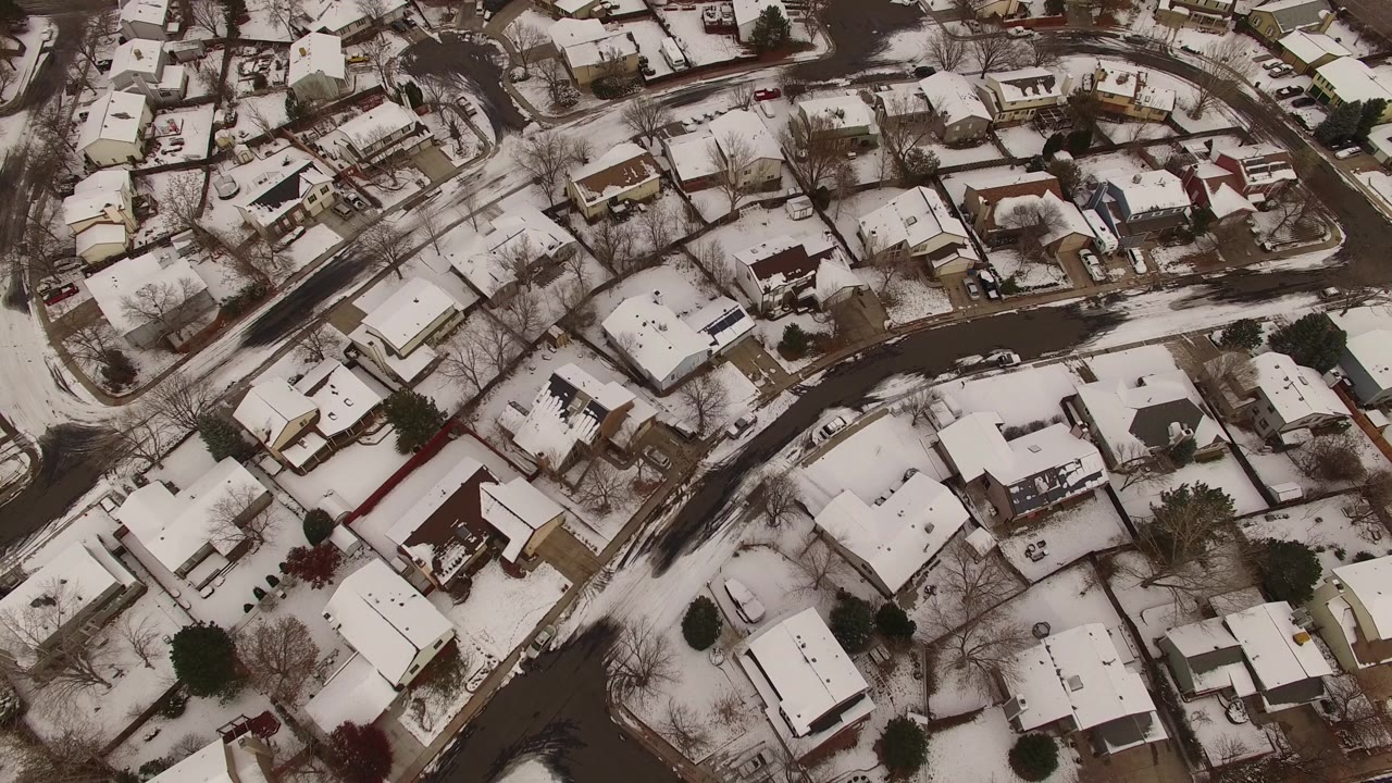 Aerial frame over Jefferson County, Colorado homes where a dark curved road cuts through snow-covered roofs, yards, and leafless trees, with no visible people.