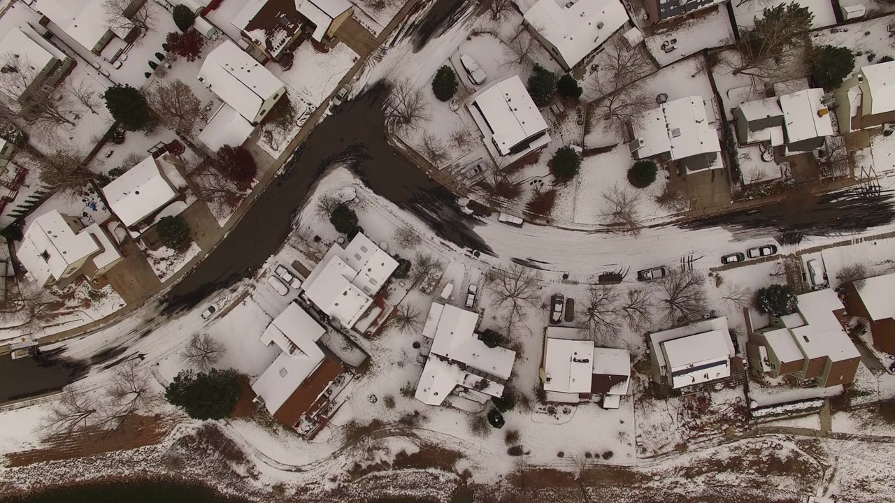 Top-down aerial view over Jefferson County, Colorado houses, snow-covered roofs, dark curving roads, parked cars, bare trees, and no visible people.