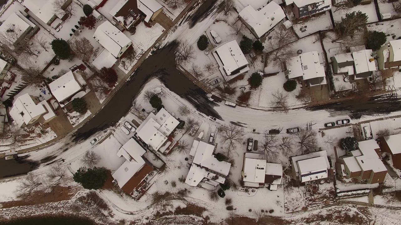 Aerial overhead frame of Jefferson County homes with white roofs, a dark curved road, driveways, parked vehicles, leafless trees, and no visible people.