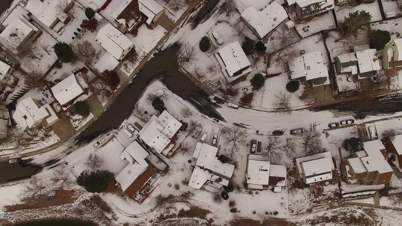 Aerial frame showing white rooftops, patchy snow in yards, a winding residential street, and a dark channel along the left side in Jefferson County, Colorado, with no visible people.