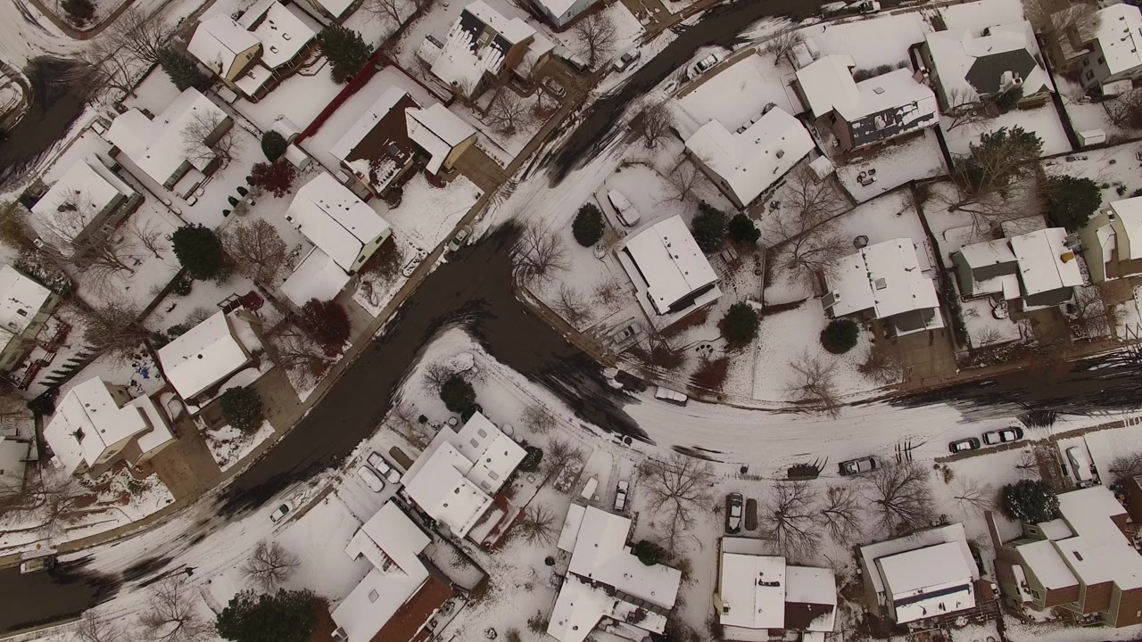Aerial frame over Jefferson County, Colorado homes, showing white rooftops, curving dark pavement, winter yards, parked vehicles, and no visible people.