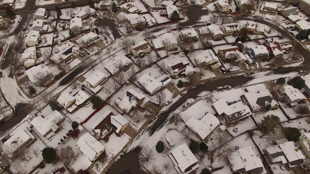 High aerial frame over Jefferson County, Colorado showing clustered homes, white yards, dark road surfaces, winter trees, and no visible people.
