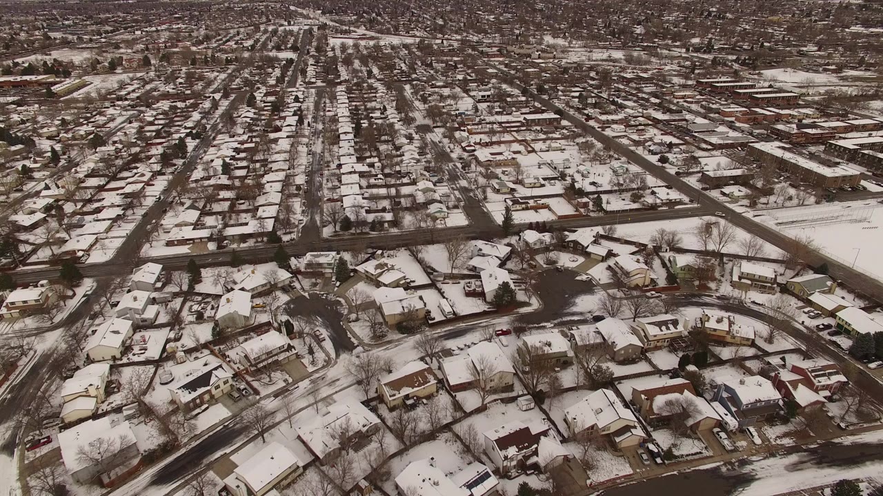 Aerial view over a snow-covered residential neighborhood in Jefferson County, Colorado, with rooftops, dark streets, bare trees, and no visible people.