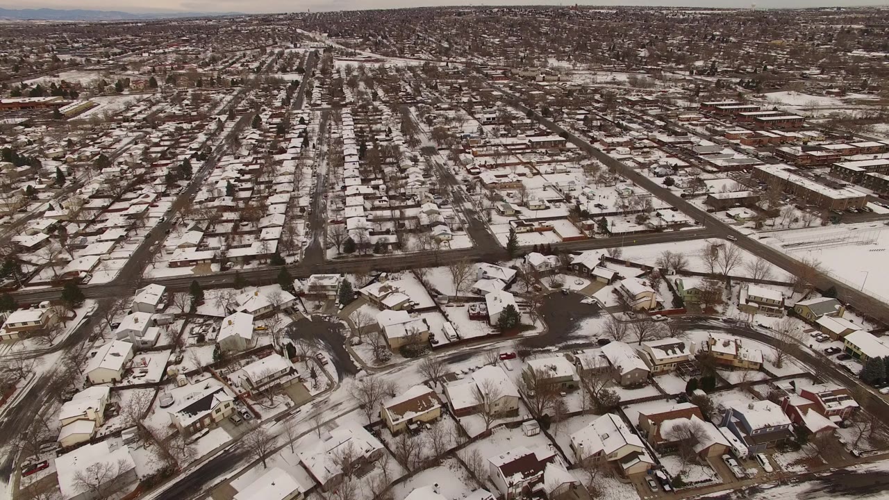 Wide aerial over snowy houses, roads, and open lots.