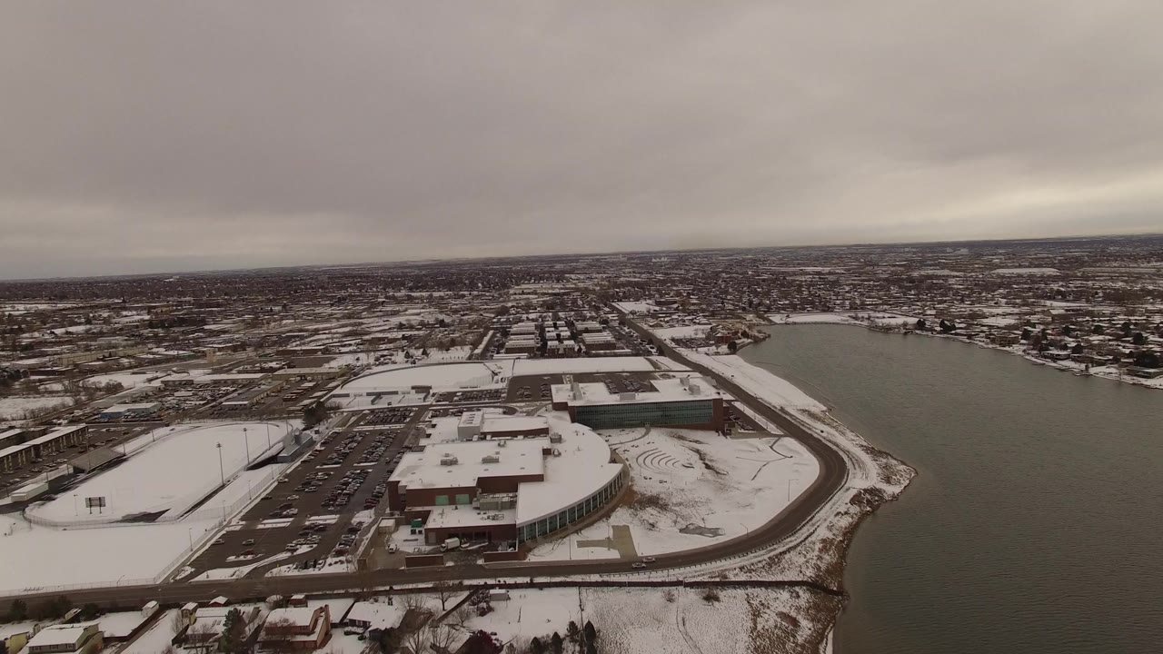 High aerial view in Jefferson County, Colorado with low buildings, dark parking lots, snowy roads, and the lake occupying the right side of the frame, with no visible people.