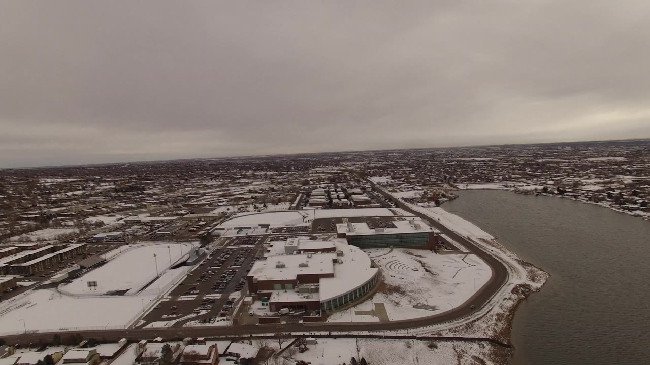Snowy aerial view over a lake, roads, parking lots, houses, and large buildings under gray clouds.