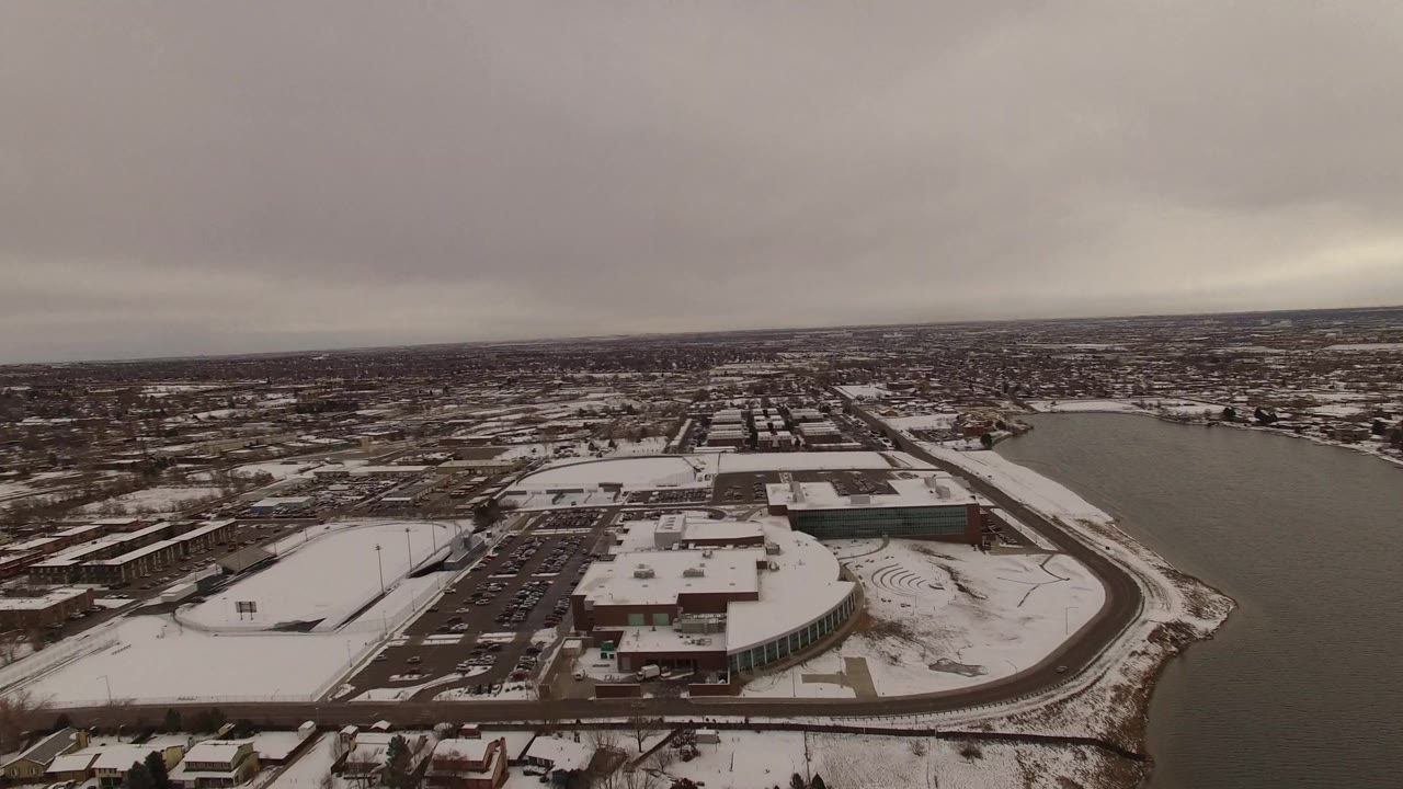 High aerial view in Jefferson County, Colorado, with the lake curving along the right side, snow on roofs and streets, clustered buildings at center, and no visible people.