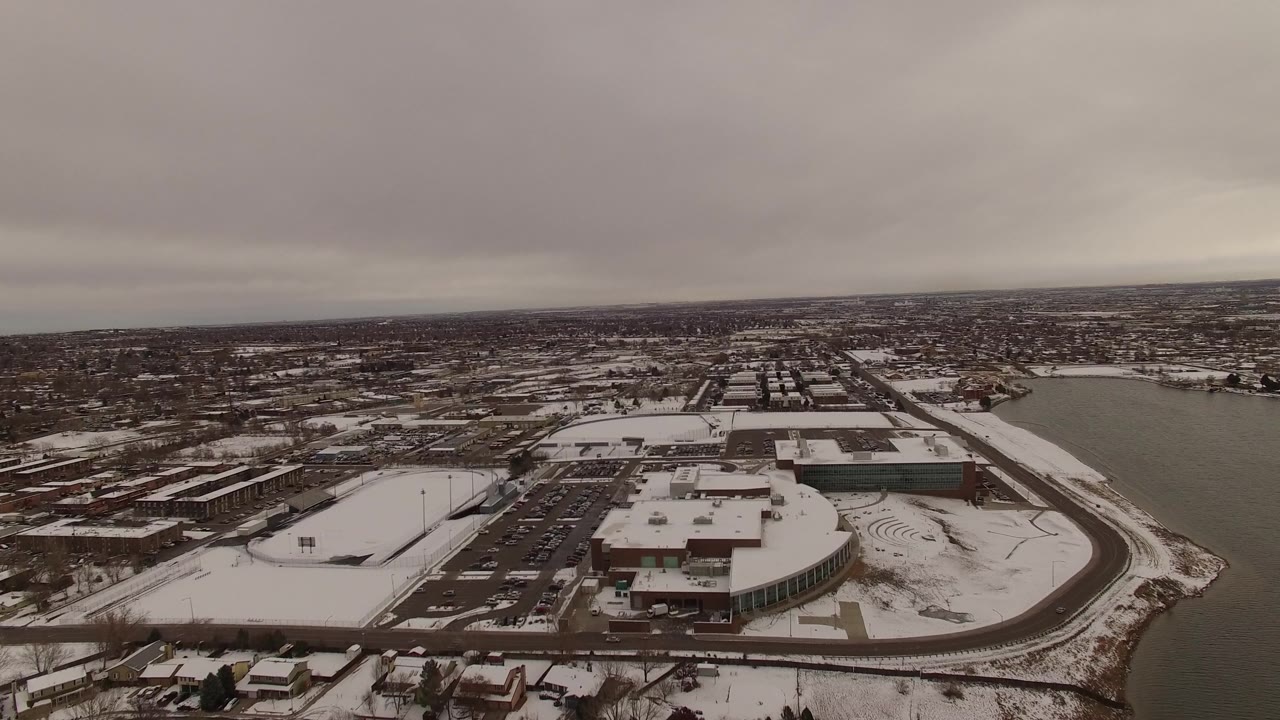 Snow-covered suburban and commercial area beside a lake with gray sky and distant city blocks.