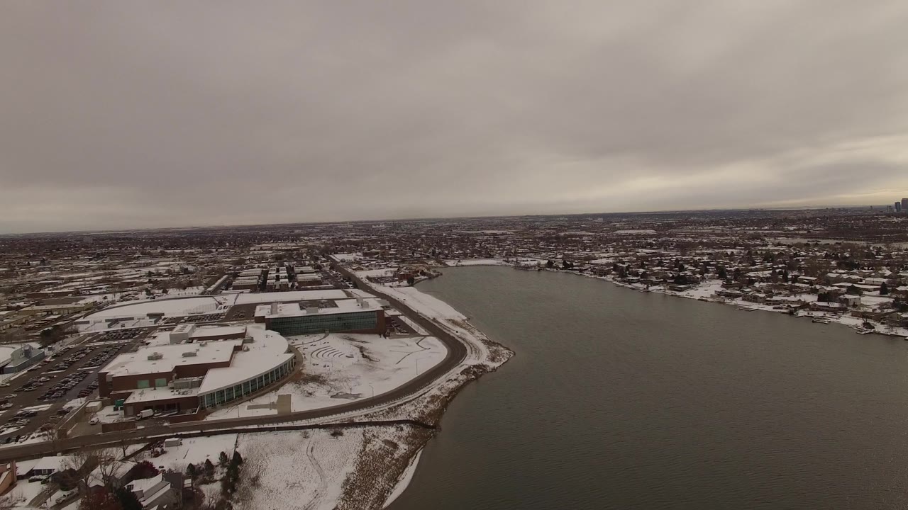Aerial frame over Jefferson, Colorado, with the waterfront building lower left, broad dark water through the center, housing across the shore, and no visible people.