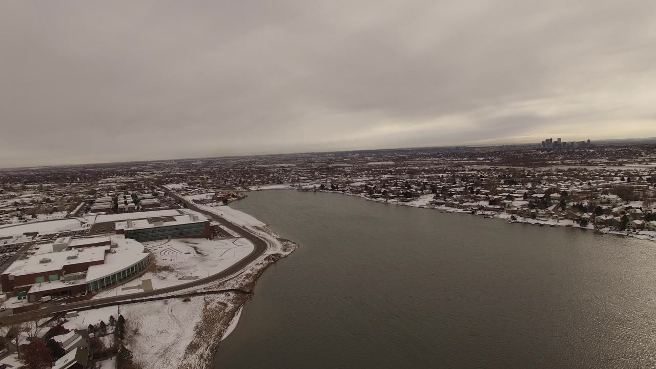 Jefferson, Colorado aerial frame with the near waterfront building lower left, broad dark water through the center, snow-dusted homes beyond, and no visible people.