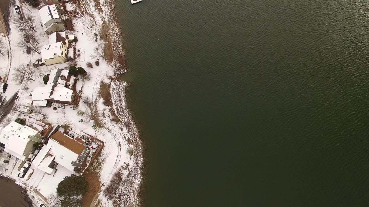 Aerial frame in Jefferson County, Colorado showing snow on rooftops and yards along a curved lake edge, with rippled dark water and no visible people.