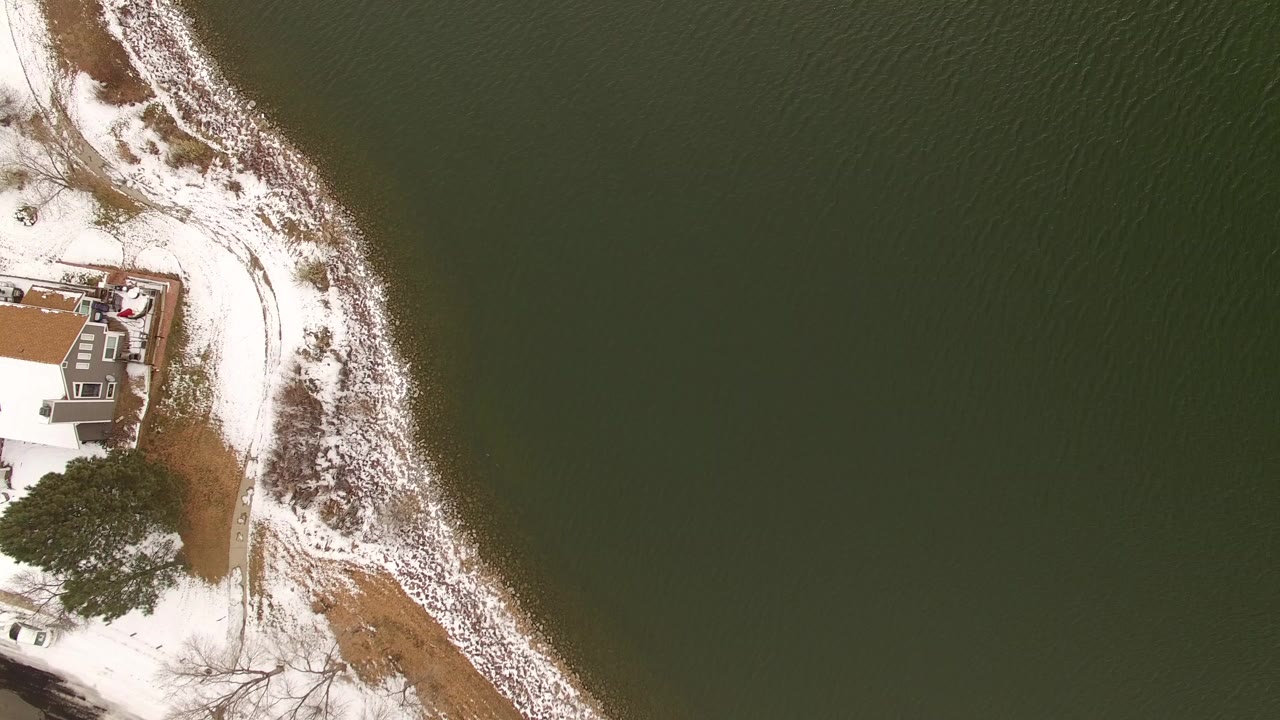 Overhead view of a snowy shoreline, dark green water, and part of a waterfront house.