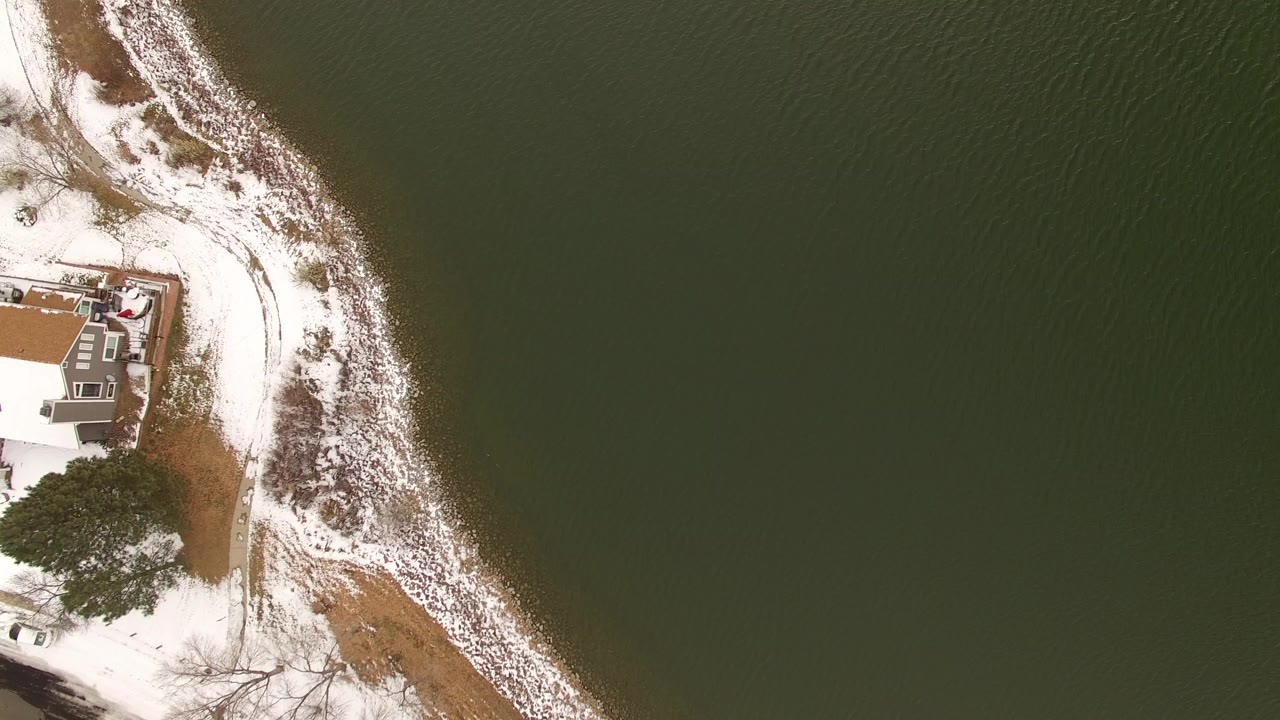Top-down frame in Jefferson County, Colorado with the waterfront house more centered near the left side, a curved strip of snow along the shore, rippled water, and no visible people.