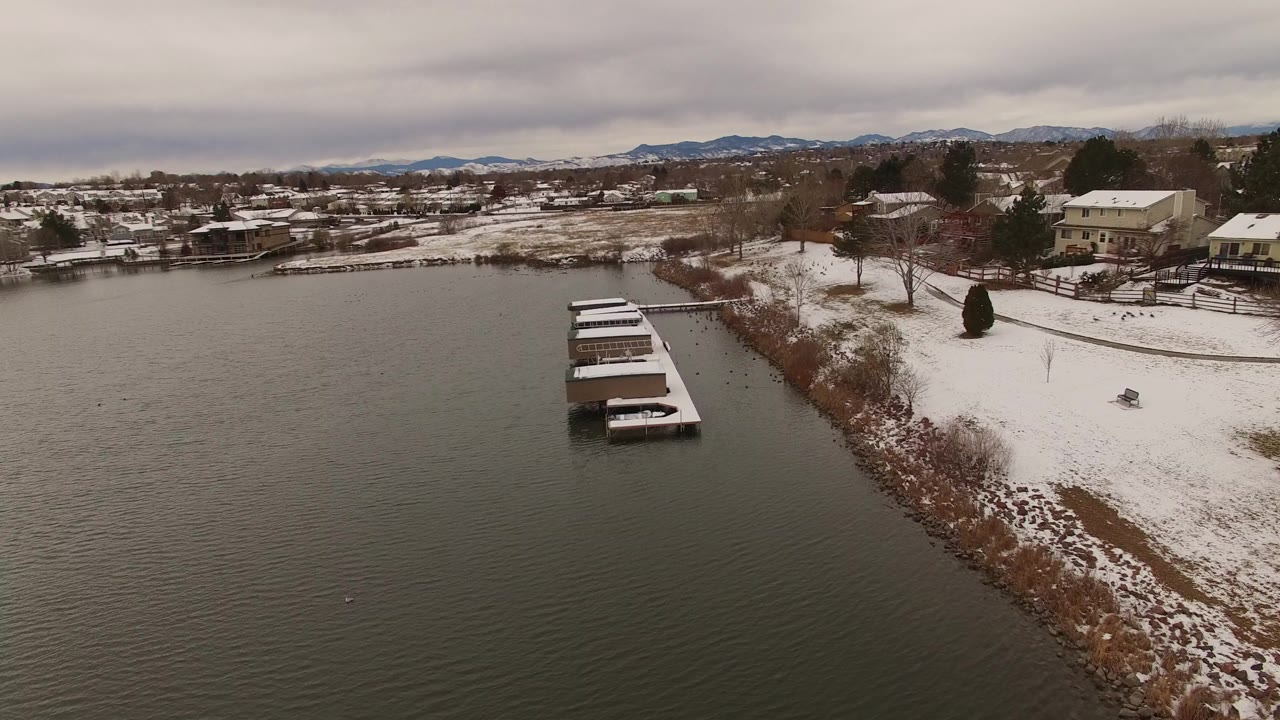 Snowy lake shoreline with covered docks and homes under gray clouds.