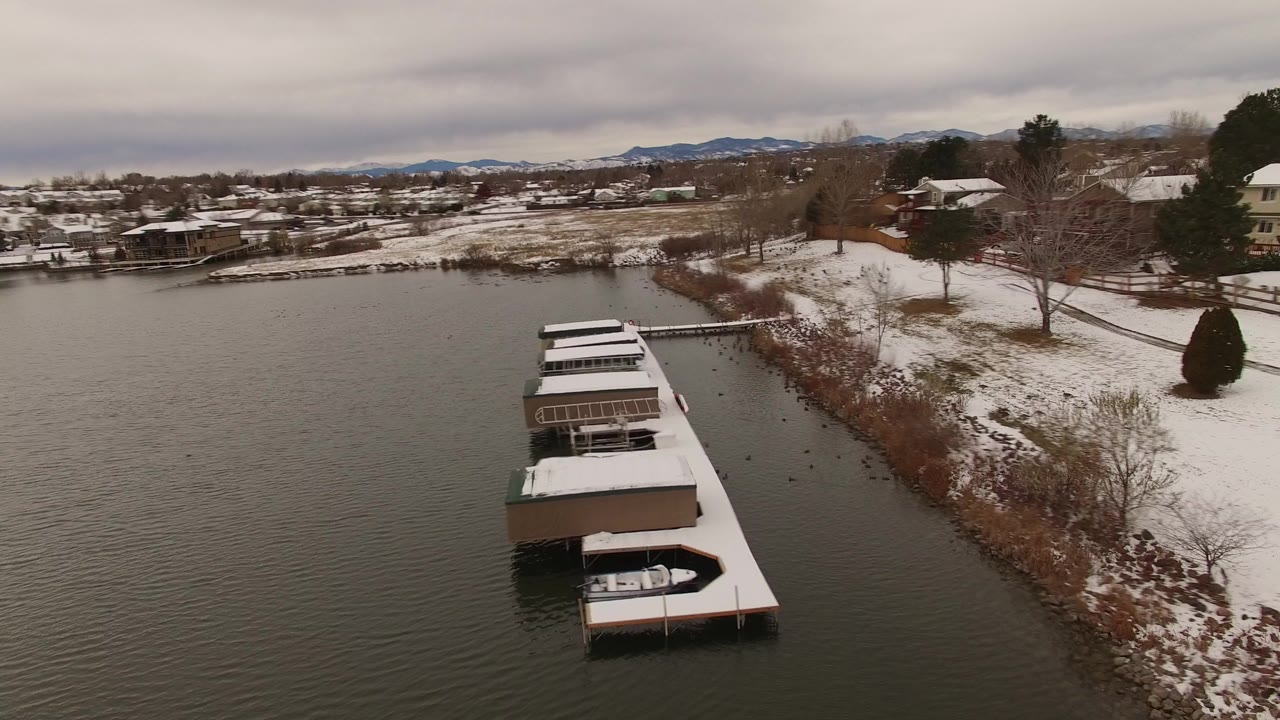 Elevated view in Jefferson County, Colorado of snow-covered boat docks along dark lake water, with a snowy shoreline, houses, bare trees, distant low mountains, and no visible people.