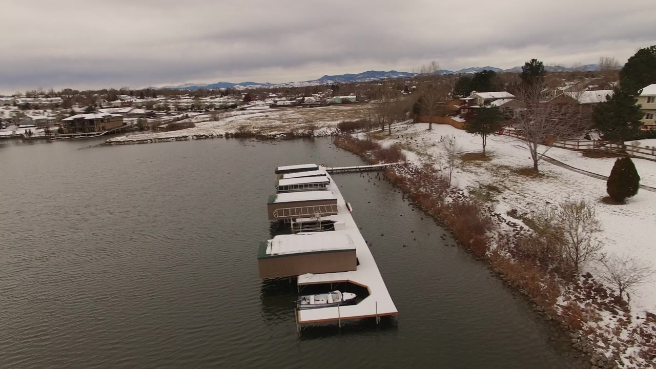 Aerial frame in Jefferson County, Colorado looking over white dock roofs and rippled lake water toward snowy residential yards, leafless trees, low mountains, and no visible people.
