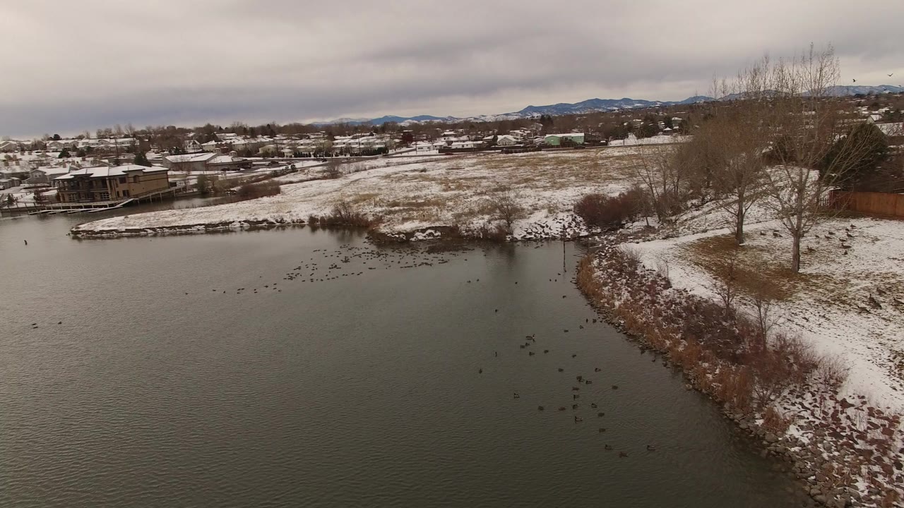Jefferson County, Colorado winter lake view with open water in the foreground, a curved snowy point at right, distant homes, low hills, and no visible people.