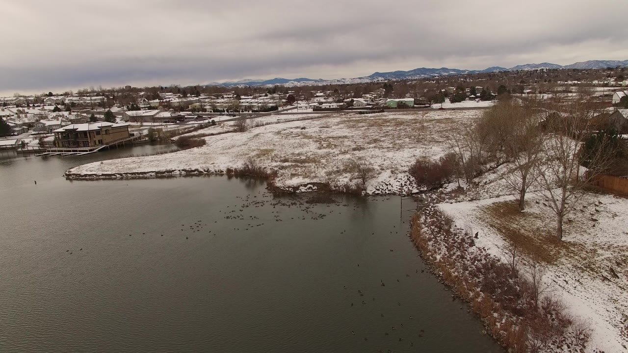 Aerial view in Jefferson County, Colorado showing dark lake water, a snow-covered shoreline, scattered houses, bare trees, distant mountains, and no visible people.