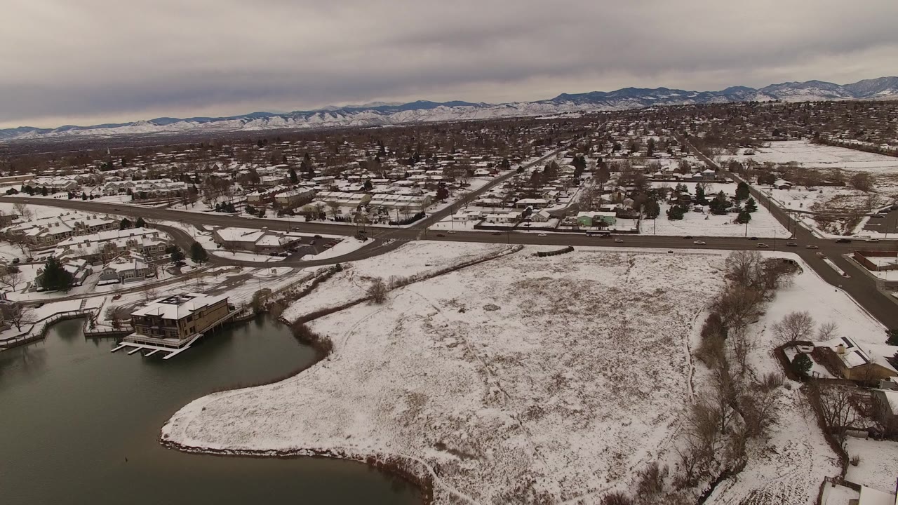 Snowy suburban aerial with lake edge, docks, roads, houses, and distant mountains.
