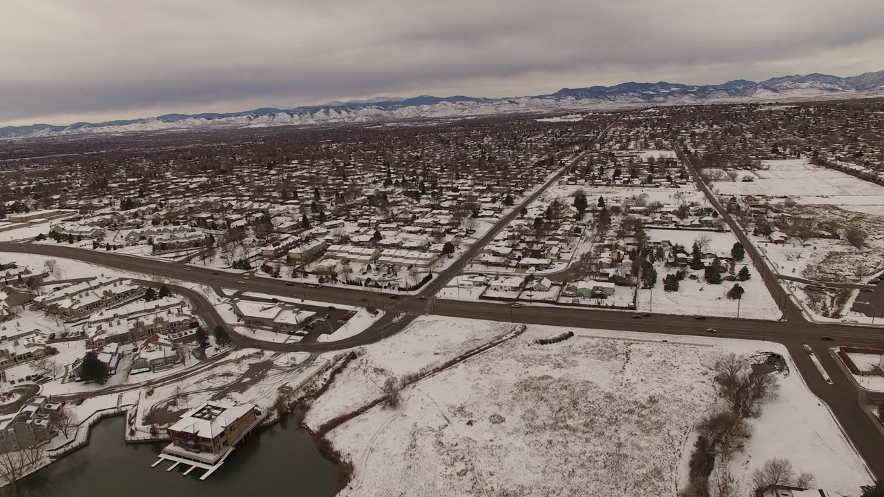 Snowy suburban aerial with roads, homes, lake edge, and distant mountains.