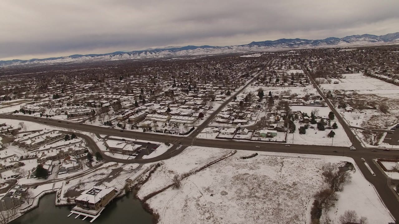 Wide aerial view over Jefferson County, Colorado, with snow-covered homes, open lots, dark roadways, a lakeside corner, distant mountains, and no visible people.