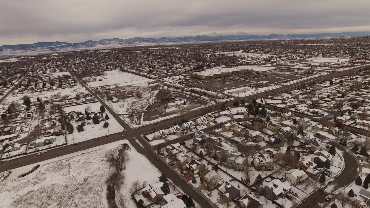 High aerial view over Jefferson County, Colorado homes, snowy yards, intersecting roads, and a low mountain horizon under gray dusk clouds, with no visible people.
