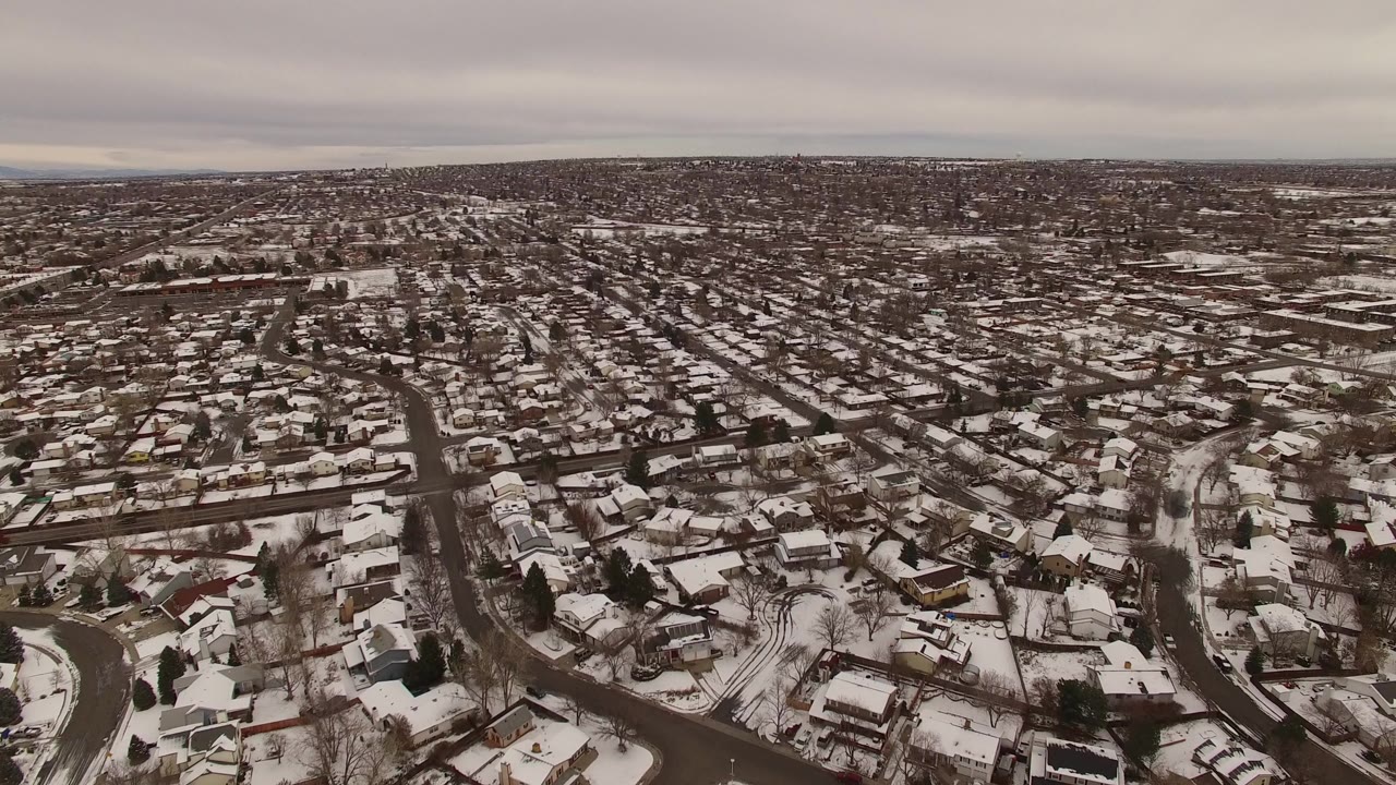 High aerial view over Jefferson County, Colorado suburbs with snow on rooftops, curved streets, bare trees, distant horizon, and no visible people.