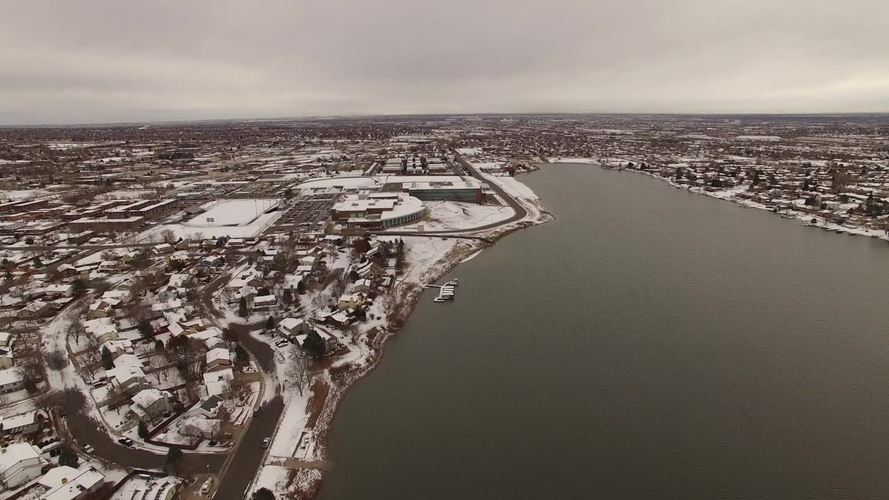 Wide aerial view of snowy lakeside neighborhoods beside dark water under gray clouds.